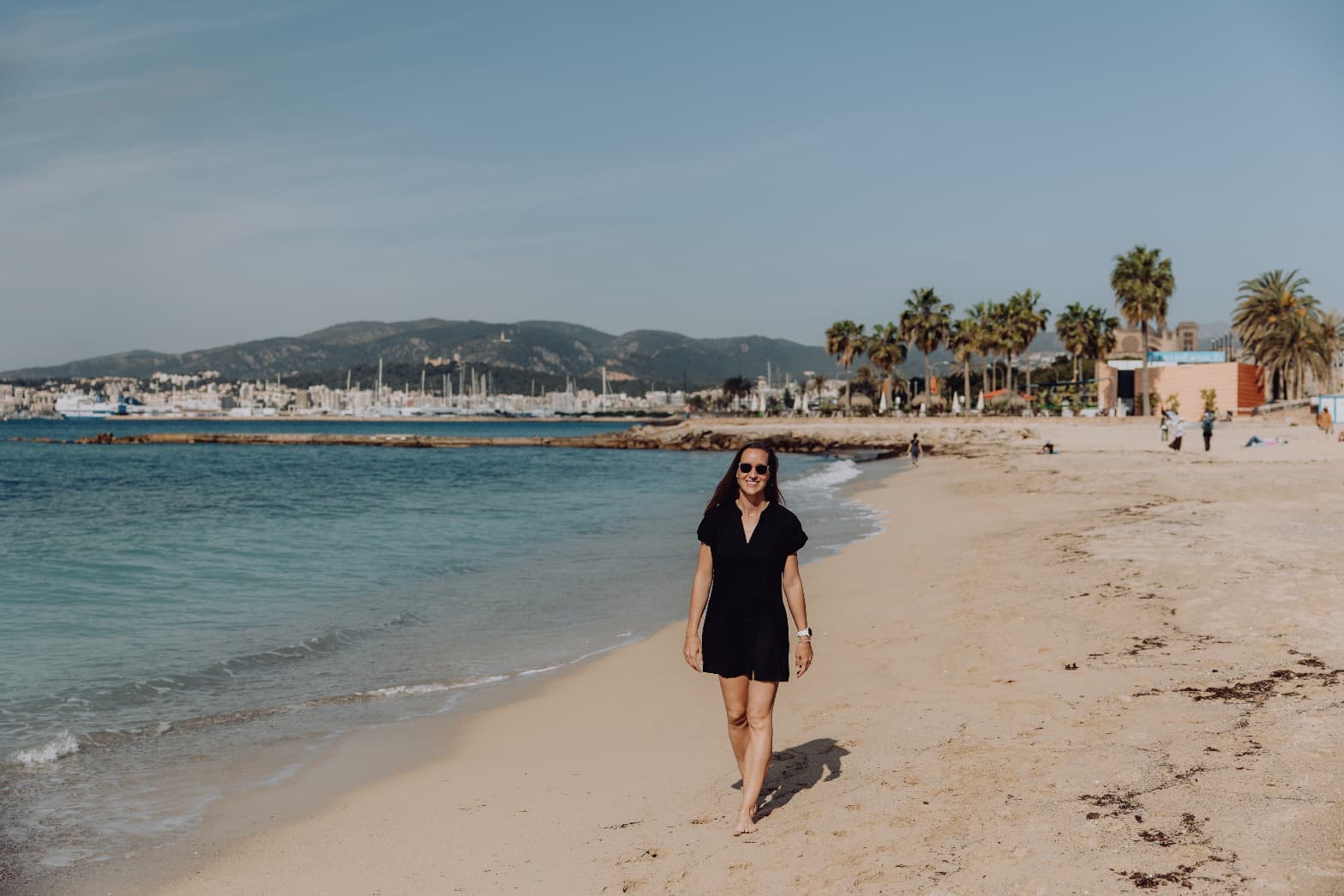 Sofie walking on a sunny beach in Mallorca with marina and mountains in the background
